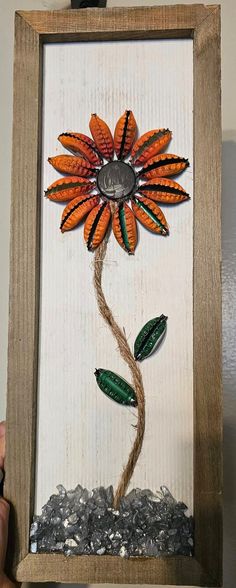 an orange flower with green leaves is in a wooden frame on the wall next to gravel