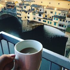 a person holding a cup of coffee in front of a bridge and buildings on the other side