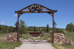 an iron and wood gate with flowers in the center on a dirt path surrounded by grass