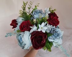 a woman's hand holding a bouquet of red, blue and white flowers with greenery