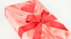 a pink gift box with a red bow and heart on it's side, wrapped in satin ribbon