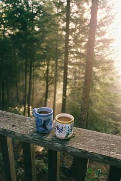 two cups of coffee sit on a wooden railing in front of the woods and trees