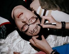 a man with glasses and writing on his face laying in bed next to a stuffed animal