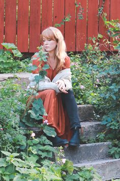 a woman sitting on steps in front of some flowers and plants with a red fence behind her