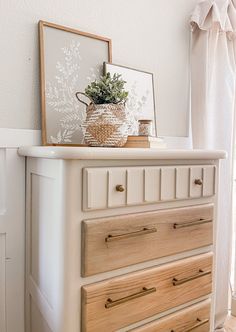 a white dresser with wooden drawers and a potted plant on top, in front of a window