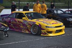 two men standing next to a yellow car in a parking lot with other cars behind them