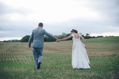 a bride and groom holding hands walking through the grass in front of an empty field