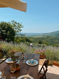 an outdoor table with plates and wine glasses on it in front of a scenic view