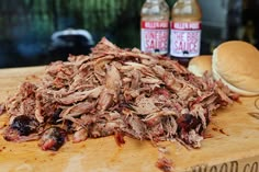 pulled pork on a cutting board next to two bottles of bbq sauce and a bun