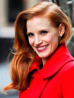 a woman with red hair is smiling and wearing a red coat on the street in front of a building