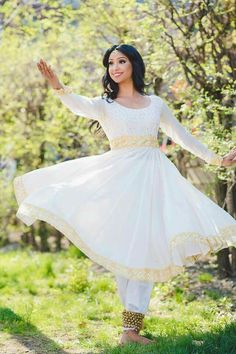 a woman in a white dress standing on grass with trees in the background and her arms outstretched