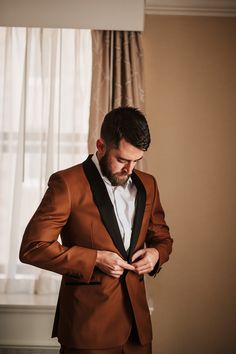 a man in a brown suit is looking down at his tie while standing next to a window