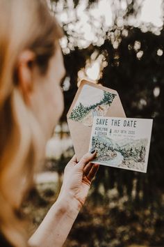 a woman holding up a save the date card in front of her face and trees