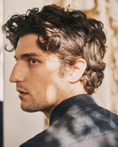 a man with curly hair wearing a leather jacket and looking off to the side while standing in front of a wall