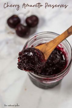 a jar filled with jam next to two cherries on a table and a wooden spoon