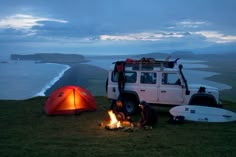 two people sitting around a campfire next to a van with a tent and surfboards