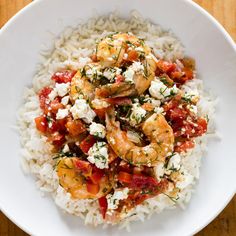 a white bowl filled with rice and shrimp on top of a bed of tomatoes, feta cheese and herbs