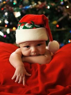 a baby wearing a santa hat on top of a red blanket next to a christmas tree