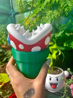 a hand holding a green and white toy in front of potted plants with faces drawn on them