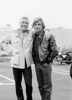 two men standing next to each other in a parking lot with their arms around each other