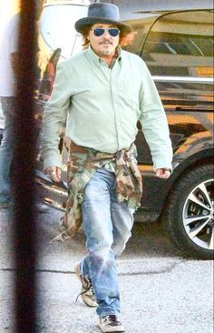 a man with a hat and sunglasses walking down the street in front of a car