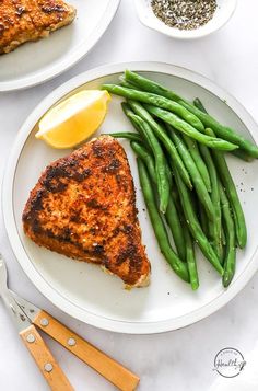 two white plates with green beans and meat on them next to some lemon wedges