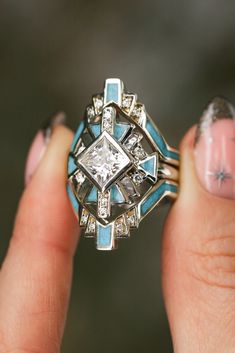 A close-up image of a woman's fingers holding a "Calliandra" design three-piece wedding ring set from Staghead Designs. This ring set features geometric Western-style design elements & a 2ct princess cut moissanite engagement ring with crushed turquoise inlays & diamond accents & is paired with two matching turquoise & diamond bands. This ring is shown in 14K white gold but can also be made in yellow or rose gold. Western Wedding Ring Stacker, Unique Turquoise Wedding Rings, Engagement Rings With Turquoise Accents, Unique Turquoise Rings, Native Wedding Rings, Southwestern Engagement Ring, Southwest Engagement Ring, Navajo Wedding Rings, Vintage Ring Sets