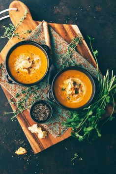 two bowls of carrot soup on a wooden cutting board with herbs and seasonings next to them