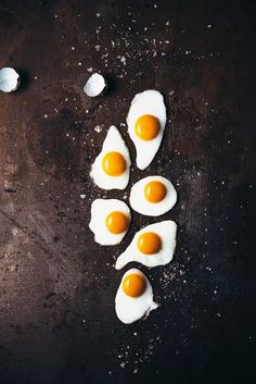 four fried eggs are arranged in the shape of five on a dark surface with salt and pepper scattered around them