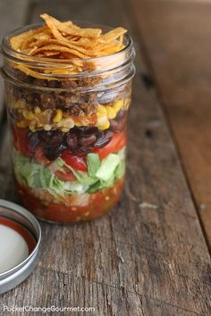 a mason jar filled with taco salad next to a container of ranch dressing on a wooden table