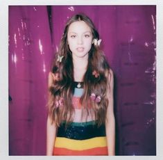 a young woman with long hair standing in front of a purple wall and wearing a colorful dress