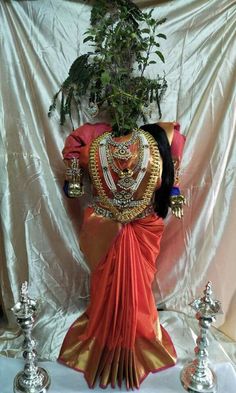 a woman dressed in an orange and gold sari standing next to two silver vases