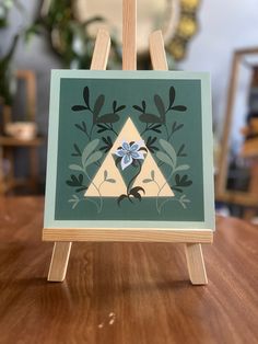 an easel with a painting on it sitting on top of a wooden table next to a potted plant