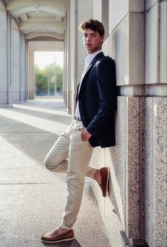 a young man leaning against a wall with his foot on the ground, wearing tan pants and a blazer