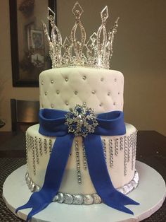 a three tiered cake with blue ribbon and tiara on top, sitting on a table