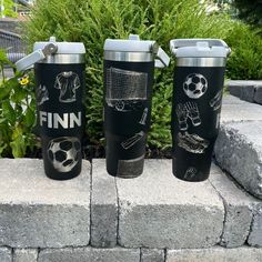 three travel mugs sitting on top of a stone wall next to plants and bushes