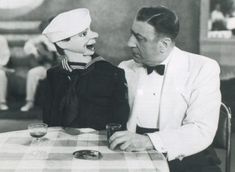 an old black and white photo of two people sitting at a table talking to each other