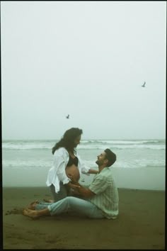 two people sitting on the beach with birds flying in the sky above them and one person holding another persons hand