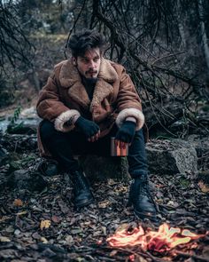 a man sitting in the woods next to a campfire with his hands on his knees