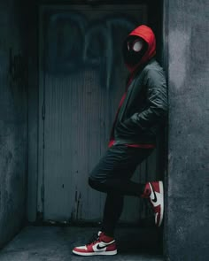 a man in a red hoodie and black jacket leaning against a wall with his foot on the ground