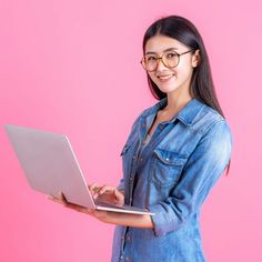 a woman wearing glasses is holding a laptop computer and smiling at the camera while standing in front of a pink background