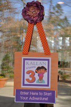 a purple and orange sign hanging from the side of a window with a flower on it