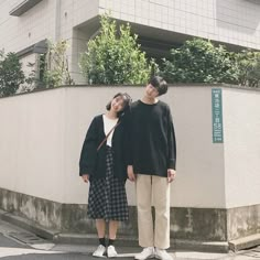 two people standing next to each other in front of a building with plants on it