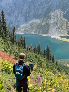 a woman with a backpack standing on top of a hill looking at a lake in the mountains