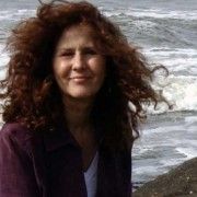 a woman with long red hair standing next to the ocean and smiling at the camera