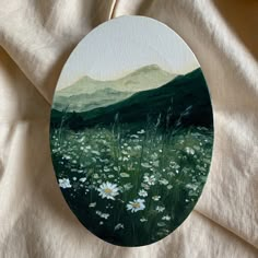 an oval painting with white flowers in the foreground and green mountains in the background