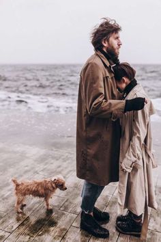 a man and woman standing on a pier next to the ocean with a dog in front of them