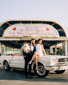 a man and woman sitting on the back of a white car in front of a sign