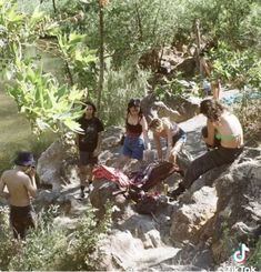 several people are standing on the rocks near water and trees, while one person is holding an object in his hand