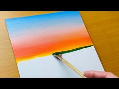 someone is painting an orange and blue sky with a paintbrush on the paper that they are holding